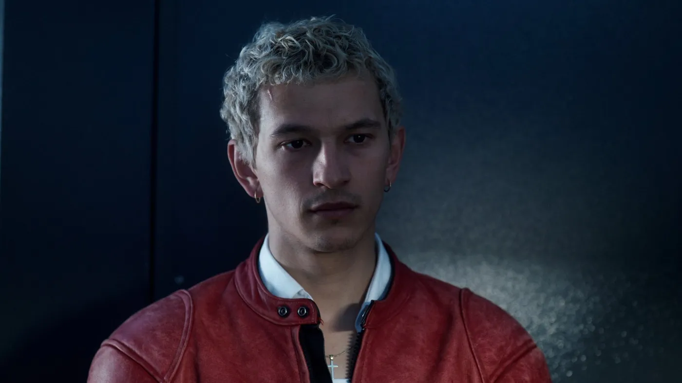 proud-7 | film.org.pl Portrait of a young man with blond curly hair wearing a red leather jacket against a dark background, looking slightly to the left.
