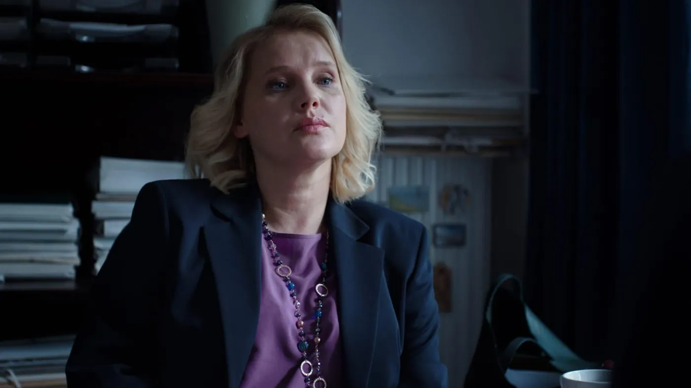 proud-8 | film.org.pl Woman with blonde hair in a navy blazer and purple blouse sits at a desk in a dim office, looking thoughtfully off to the side.