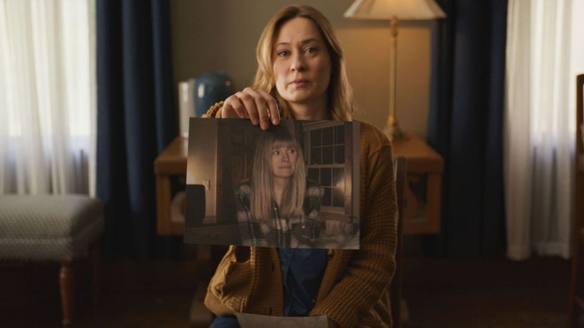Woman in a brown cardigan sits in a dim living room, holding up a photo of a younger blonde woman.