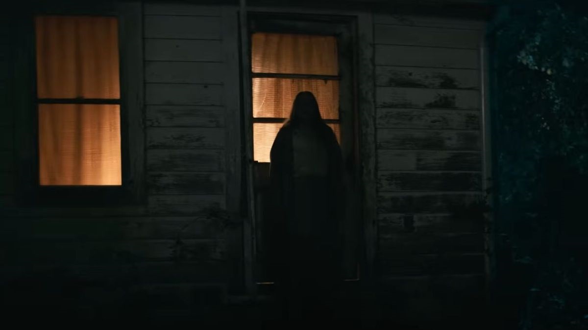 Dark night scene of a weathered house with a person standing in the doorway, illuminated by warm light from inside windowed panels.