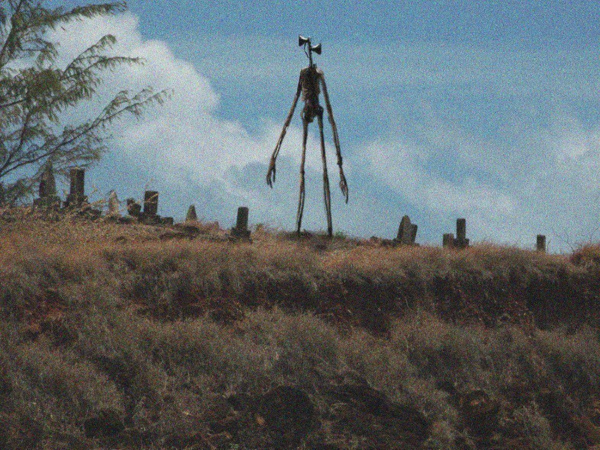 Tall skeletal sculpture with long limbs standing in a grassy hilltop cemetery amid weathered headstones under a partly cloudy sky.