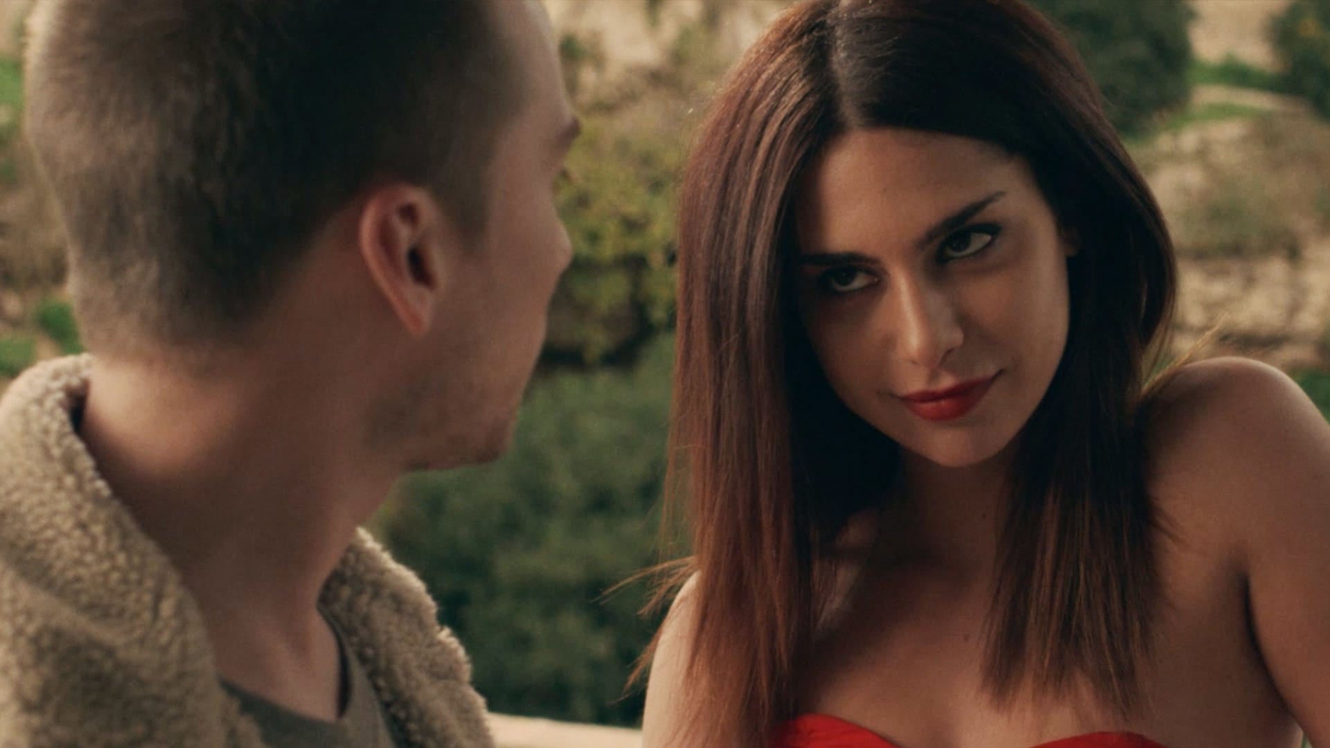spring | film.org.pl Woman with red lipstick looks sideways at a man outdoors, smiling subtly during a conversation in a sunlit setting.