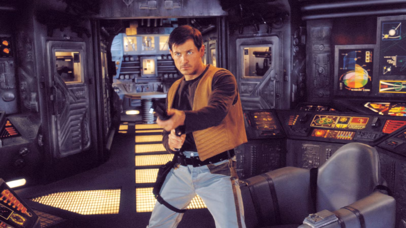 Man in a tan vest and jeans points a blaster in a high-tech spaceship cockpit with illuminated control panels nearby.