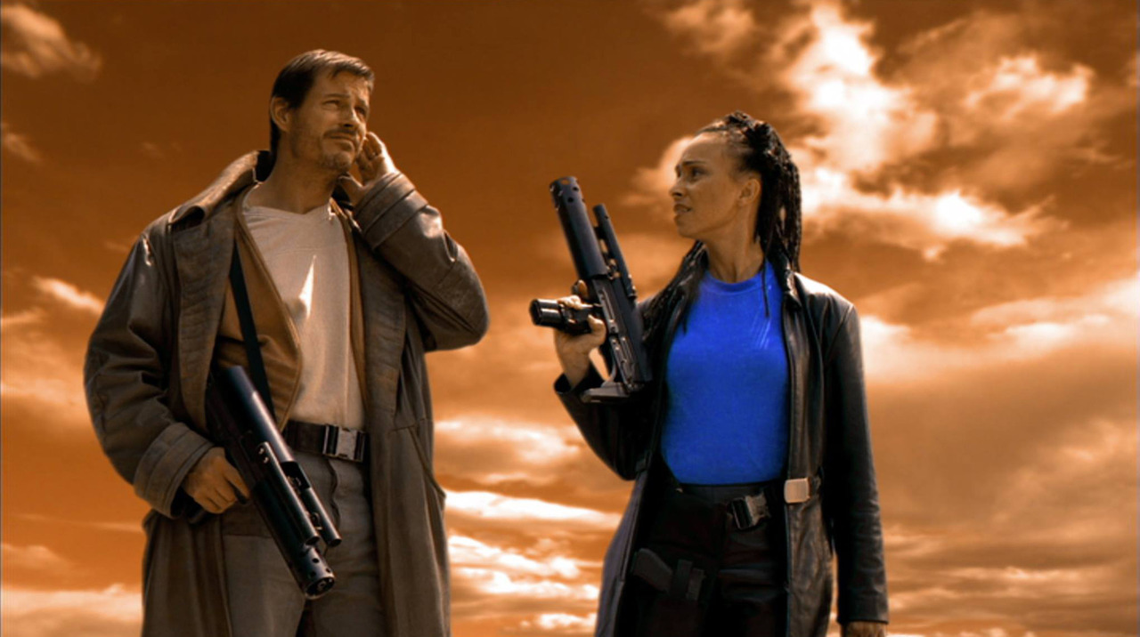 Two adults in rugged coats stand against an orange sky, each holding a large futuristic-looking gun and chatting.