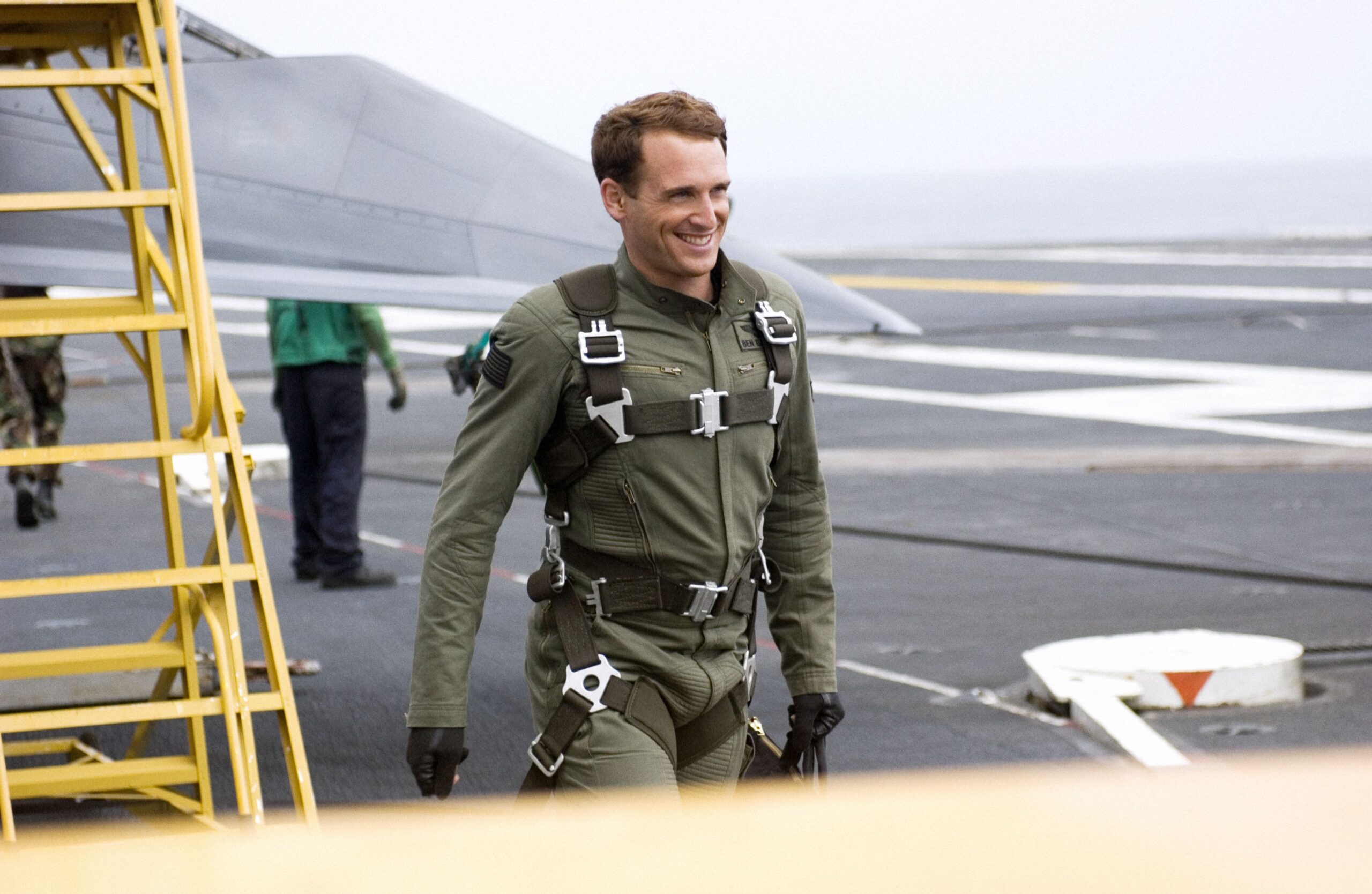 Pilot in a green flight suit with a harness walks on an aircraft carrier deck, with a yellow maintenance ladder in the foreground and a parked jet in the background.