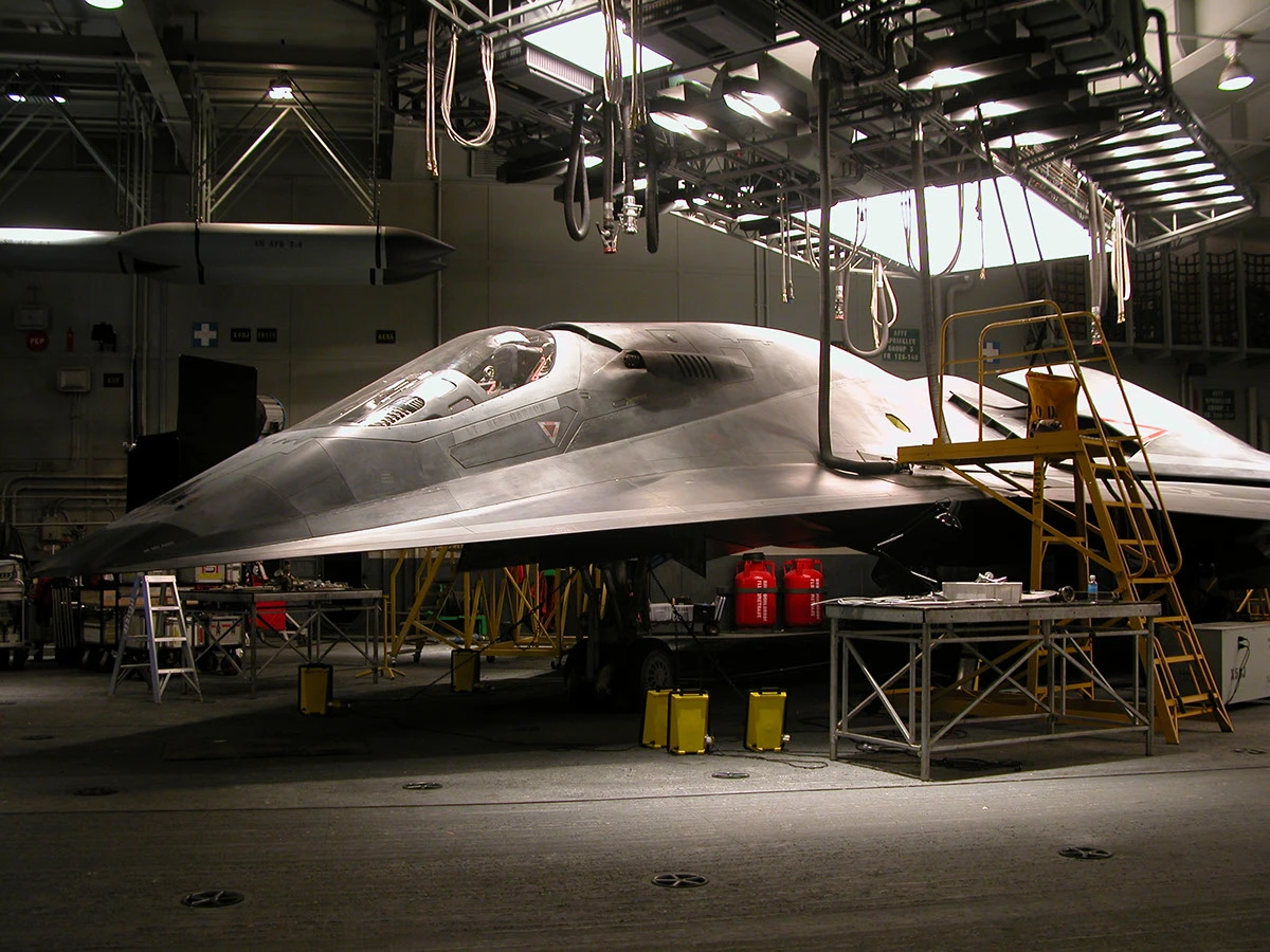 Military jet in a hangar undergoing maintenance, with scaffolding, tools, and yellow mobile steps nearby.