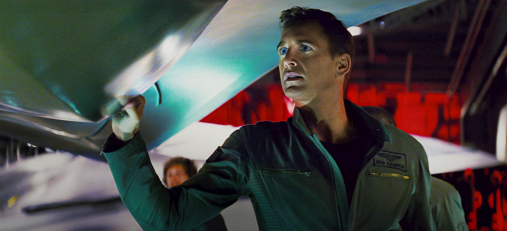 Pilot in a green flight suit inspects the wing of a jet inside a hangar with red lighting in the background, focused expression on his face.