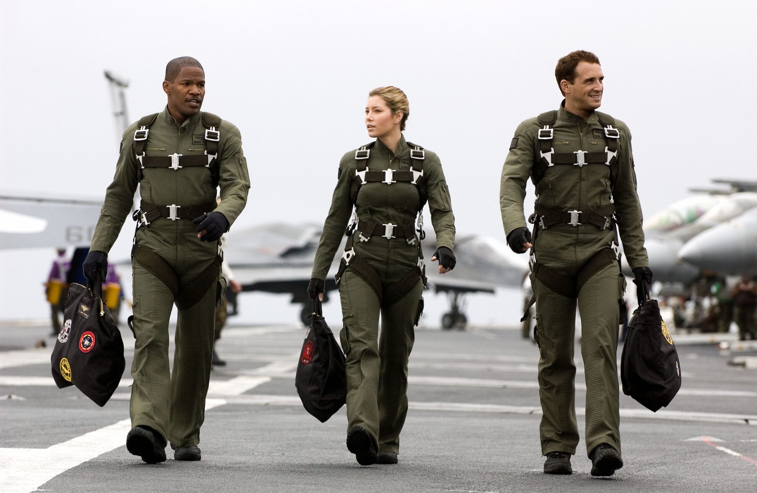 Three pilots in olive flight suits walk briskly on a tarmac, each wearing parachute harnesses and carrying black gear bags, with aircraft in the background.