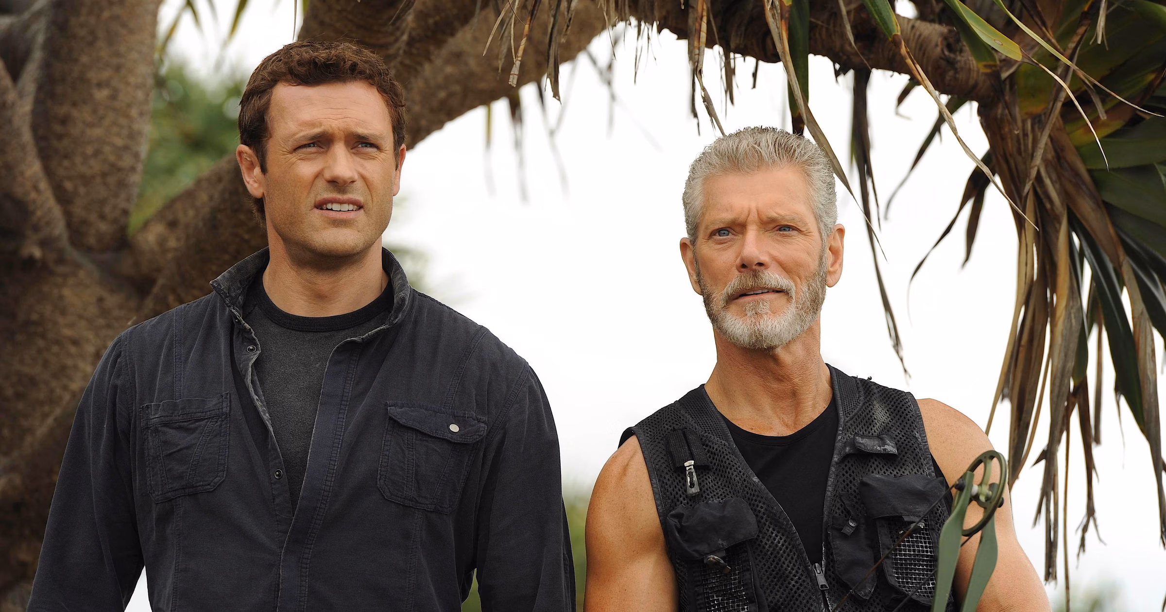 terra-nova-2 | film.org.pl Two men stand side by side outdoors under a tree; the younger man wears a dark shirt, the older man sports a gray beard and a black tactical vest with archery gear nearby.