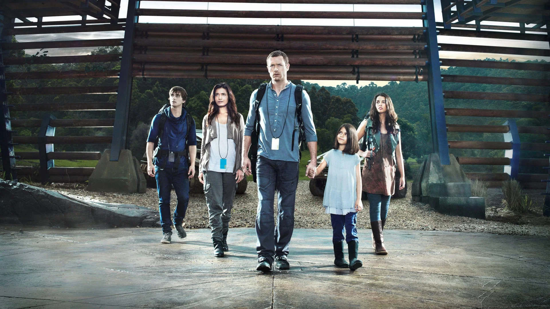terra-nova | film.org.pl A family of five with backpacks walks toward the camera under a wooden, log-backed structure in a forested outdoor area.
