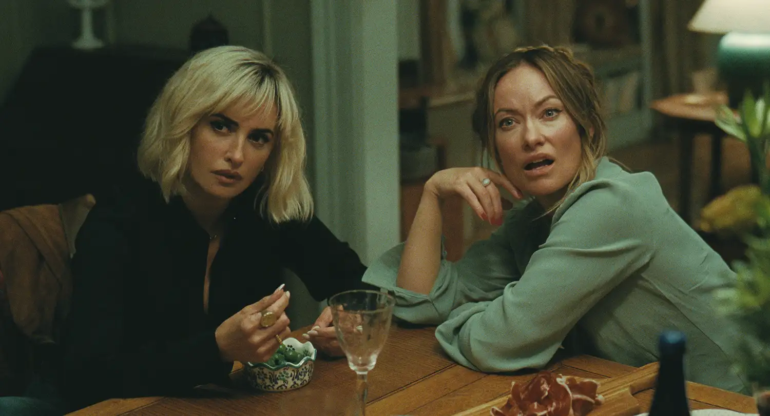 Two women sit at a wooden dining table indoors, one with blonde hair in a black outfit and the other in a light green blouse, both looking surprised or concerned.