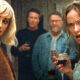 Two women at a party, facing the camera and holding drinks, with two men blurred in the background.