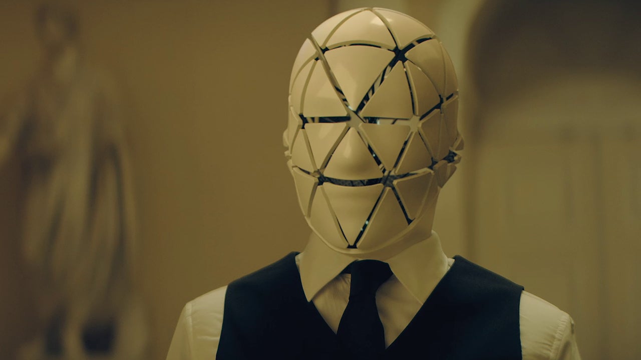 the-peripheral-peryferal | film.org.pl Person wearing a white geometric panel mask over the face, dressed in a white shirt, black vest, and tie in a dim setting.