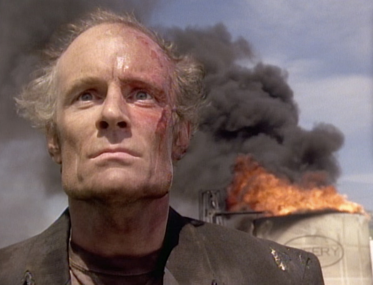 the-stand-bastion-1994-2 | film.org.pl Man with scorched face and stern expression stands in foreground as a fiery explosion burns in the background, smoke rising behind him.