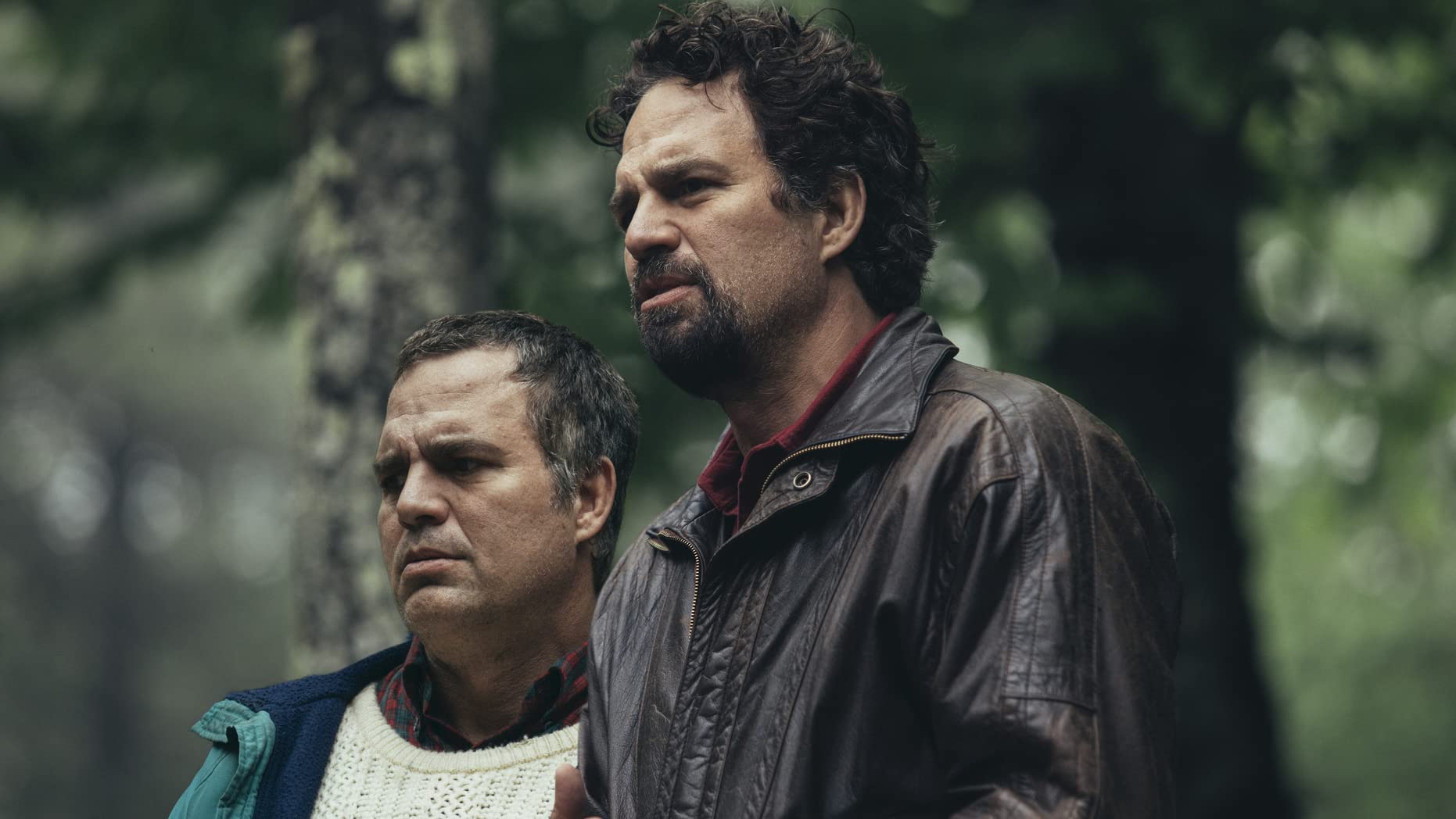 Two men stand in a forest, looking off into the distance with serious expressions.