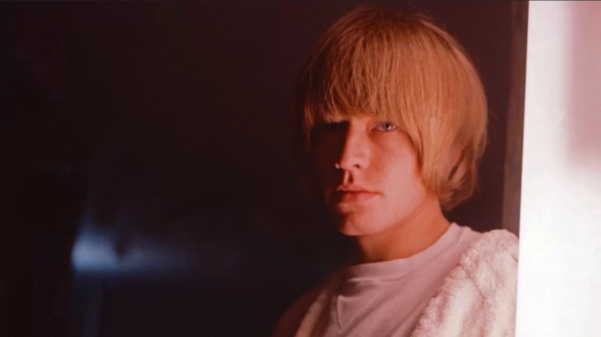 Young person with blonde hair in a white shirt, towel over shoulder, looking at the camera in a dim room.