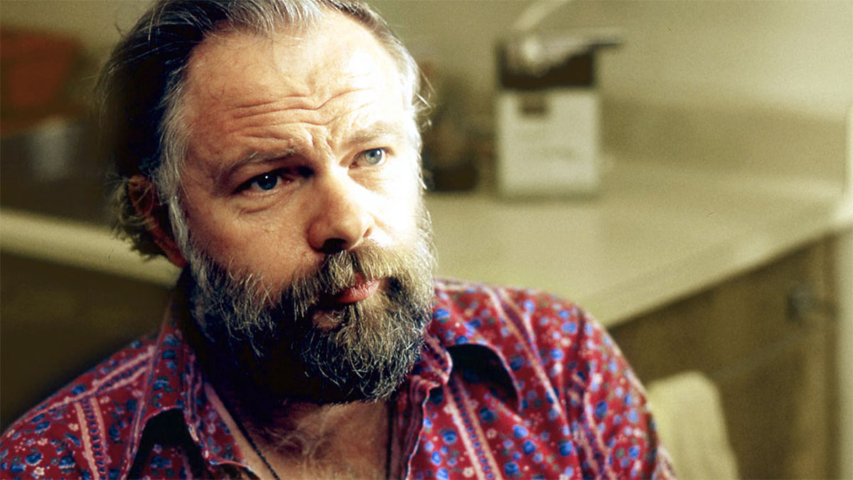 Close-up of a bearded man with a patterned red shirt, looking thoughtful in a kitchen setting.