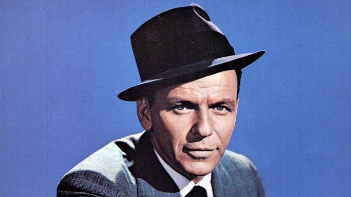 Male portrait in a dark fedora and suit against a blue background.