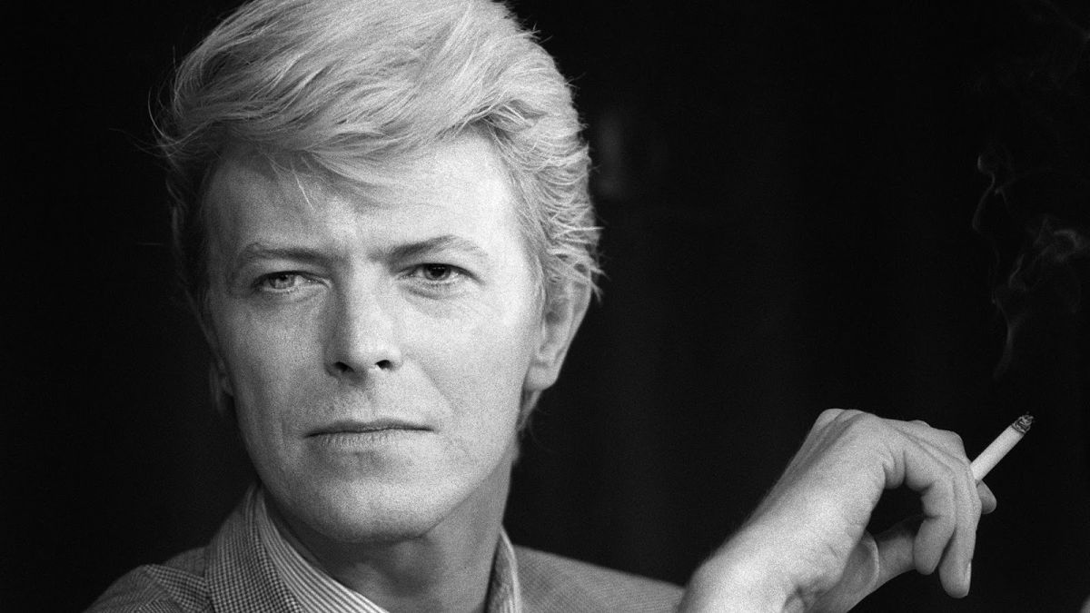 Black-and-white portrait of a middle-aged person with short light hair, smoking a cigarette and gazing at the camera.