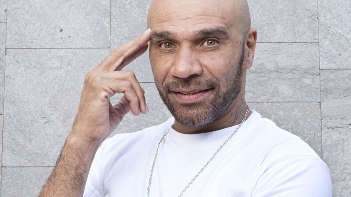 Bald man with a trimmed beard, wearing a white T-shirt, touching his temple with one finger against a gray stone wall.