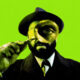 Man in a suit and fedora peers through a magnifying glass against a bright lime green background