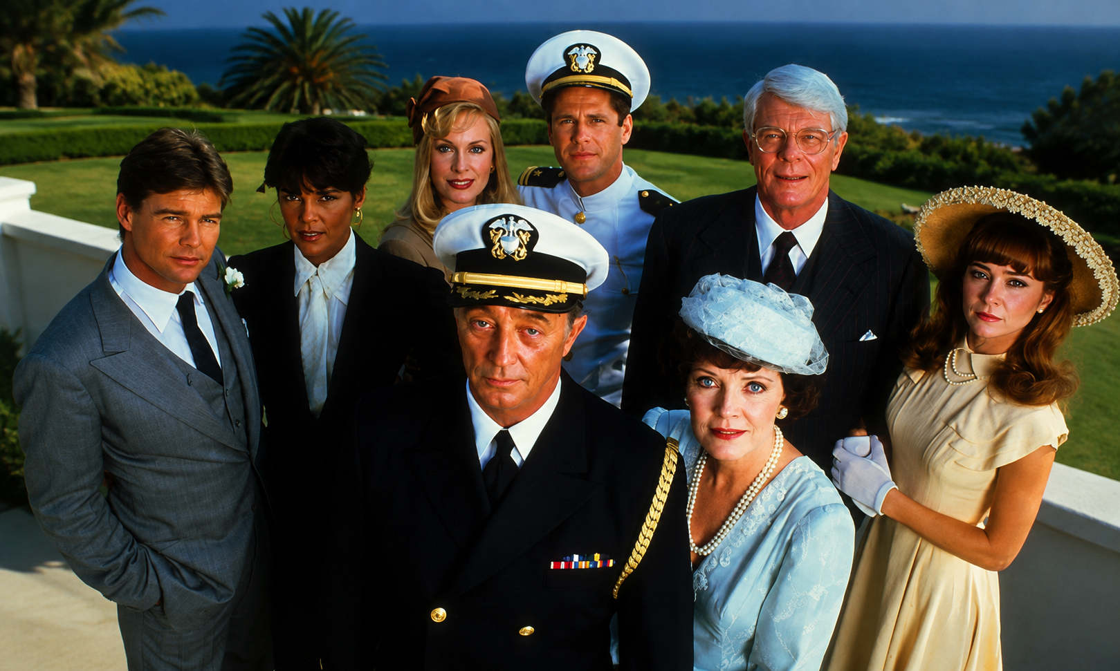 Group of formally dressed adults posing outdoors by the sea, including naval officers and a bride-like woman in a light dress, with a green lawn behind them.