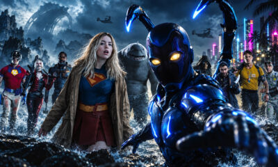 Diverse group of superheroes on a rocky shore, led by a blue armored hero reaching toward the camera as waves splash.