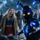 Diverse group of superheroes on a rocky shore, led by a blue armored hero reaching toward the camera as waves splash.