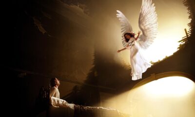 White-winged angel character in a white dress soars above a glowing circular arch while a spectator watches from the left in a dark theater.