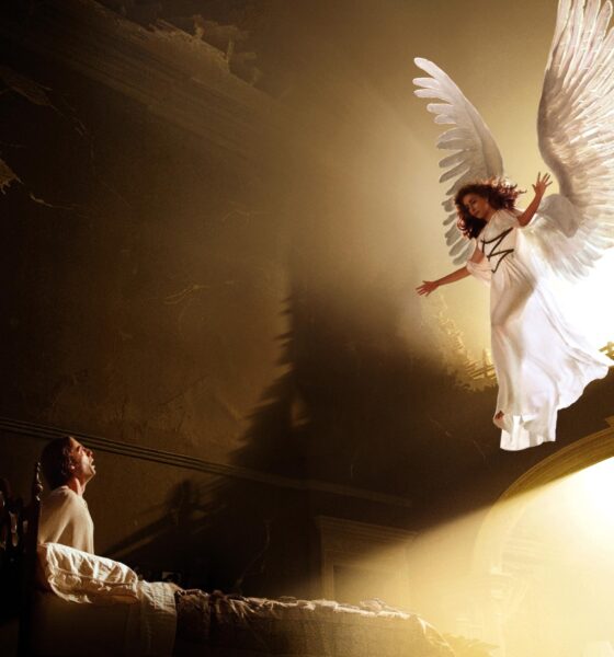 White-winged angel character in a white dress soars above a glowing circular arch while a spectator watches from the left in a dark theater.