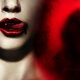 Close-up of glossy red lips with a small drop of blood, set against a red-tinged, shadowy background with a blurred screaming figure on the right.