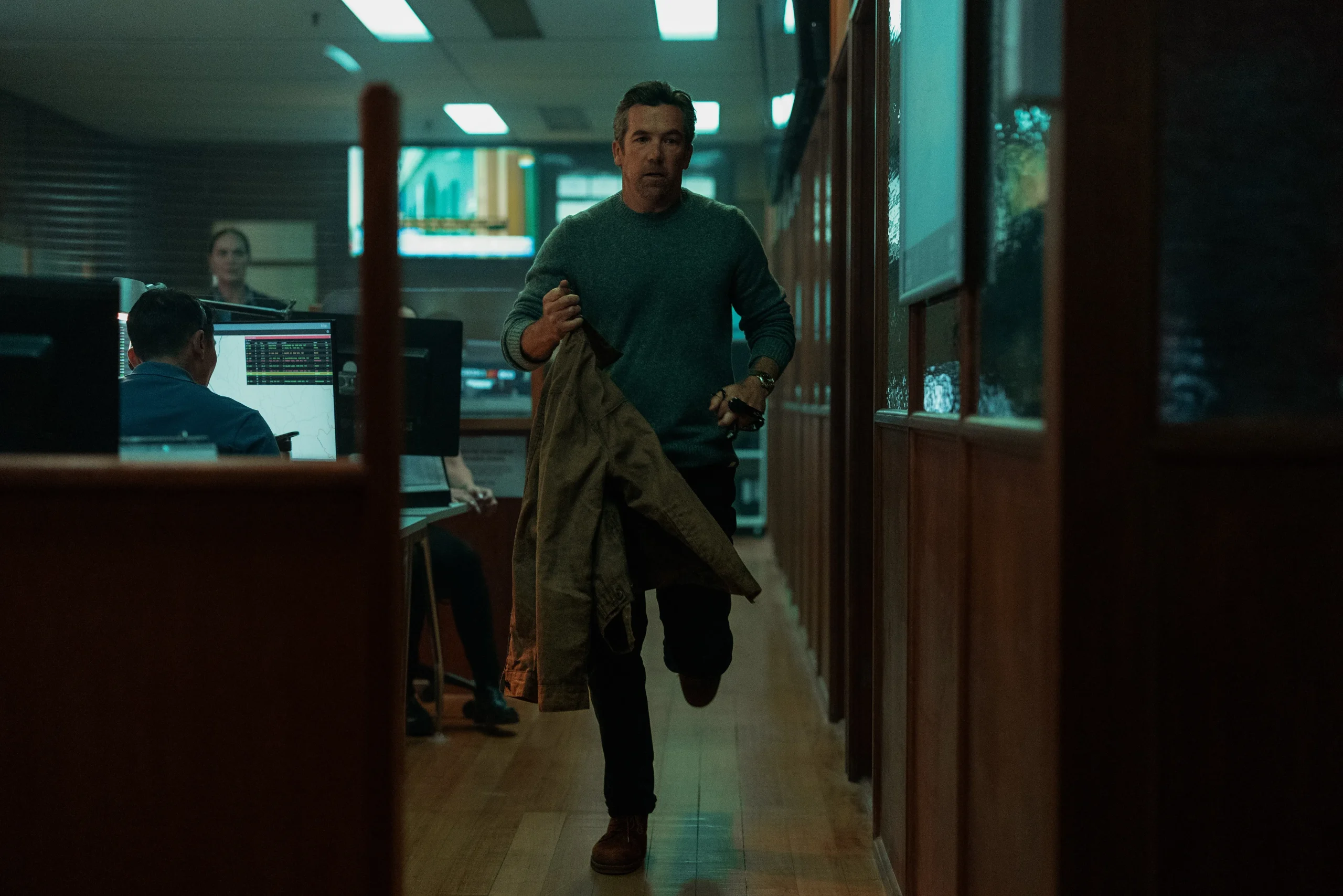 Man in a teal sweater walks down a dim office hallway, carrying a coat, as coworkers sit at computers to the left.