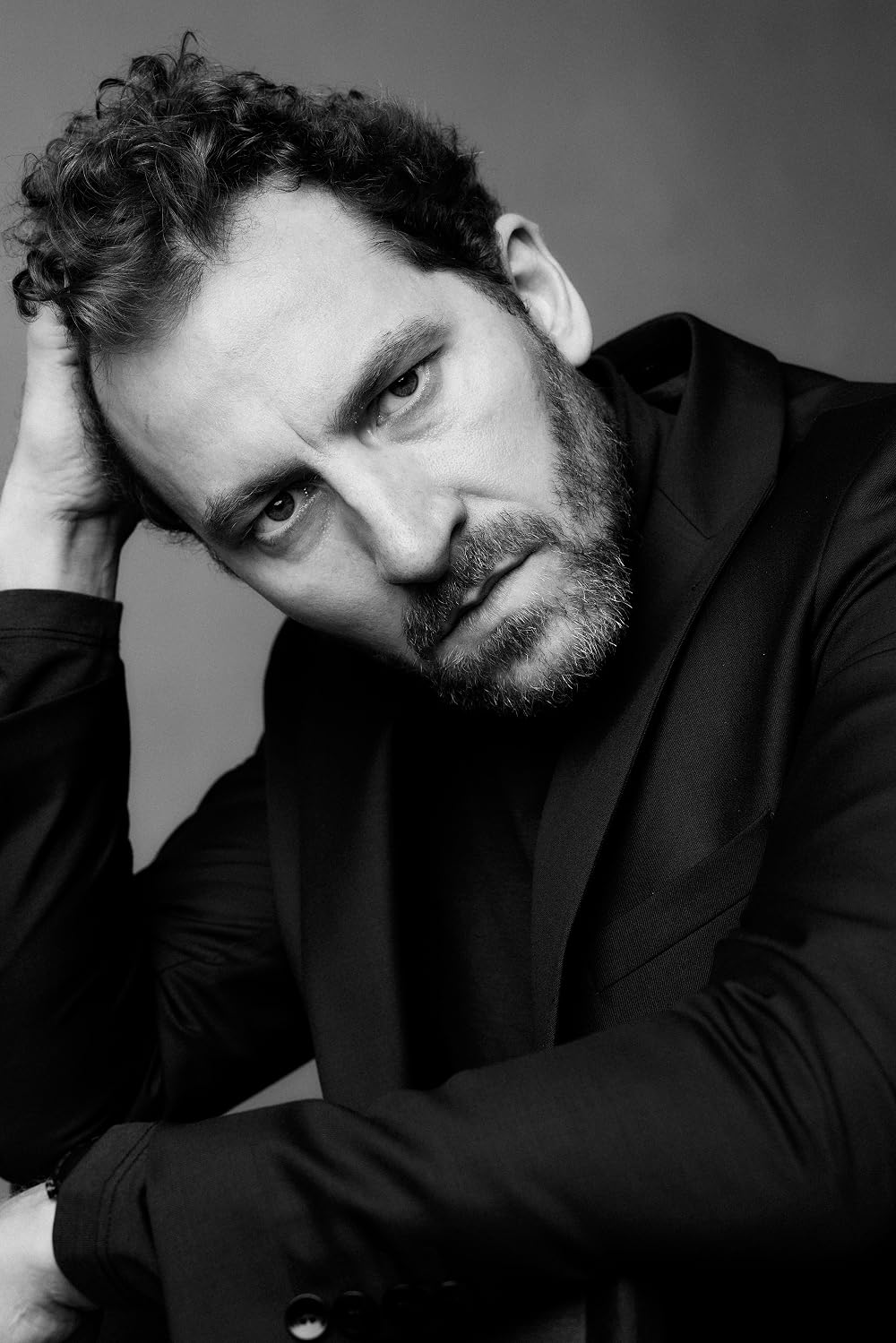 Black-and-white portrait of a bearded man in a dark blazer resting his head on his hand, looking thoughtful to the side.