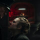 Police officer leans toward a bloodied woman in a dark, dungeon-like room with a red-lit background and a candlelit table in the distance.
