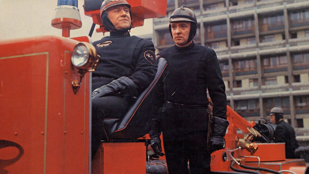 Kadr z filmu 451 stopni Fahrenheita: dwaj mężczyźni w czarnych mundurach i hełmach przy czerwonym wozie strażackim, w tle wysoki budynek. Filmy sci-fi na podstawie książek