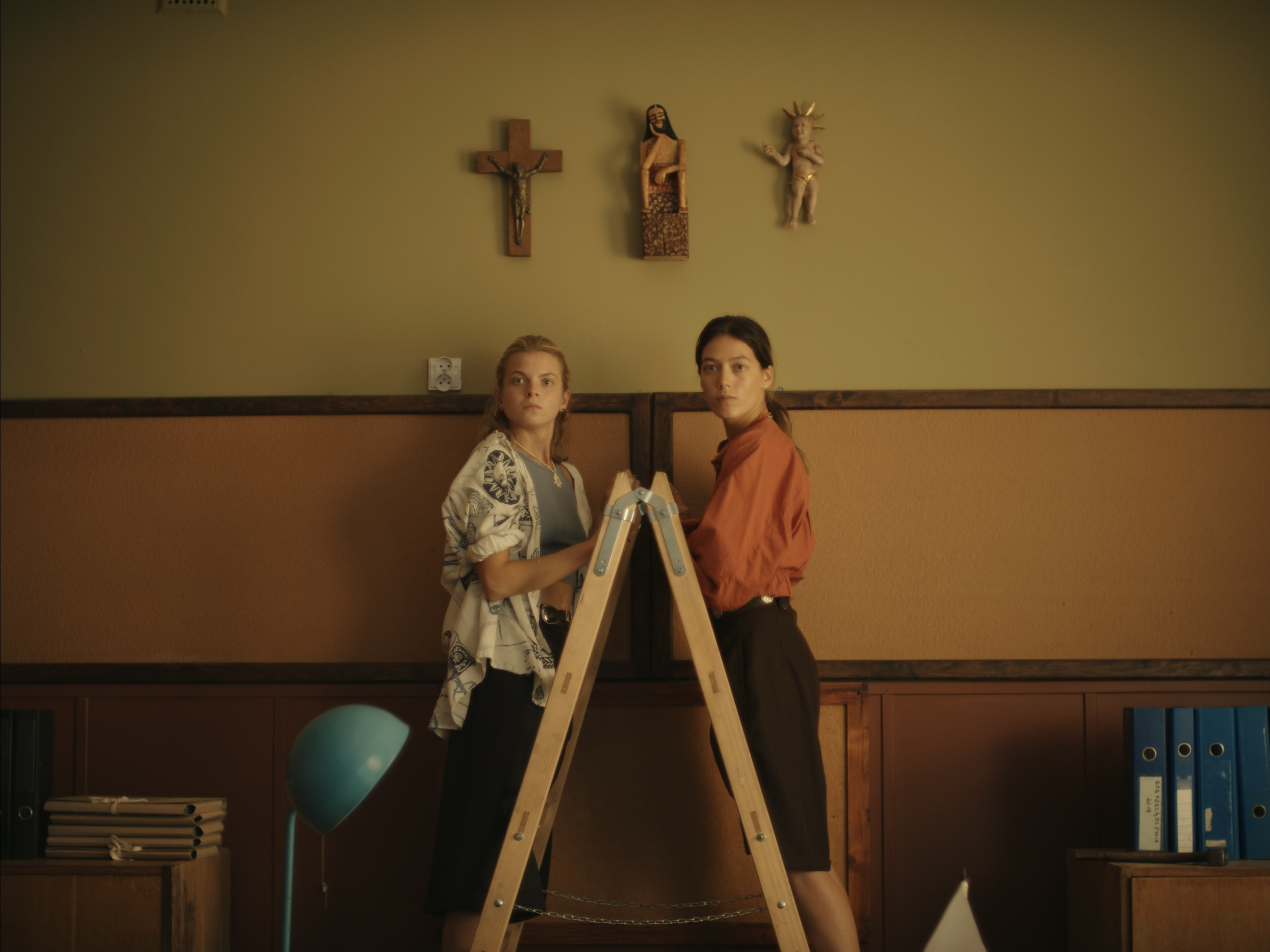 Czarna godzina | film.org.pl Two women stand on a tall ladder in an office, facing the camera with a row of religious figurines on the wall behind them.
