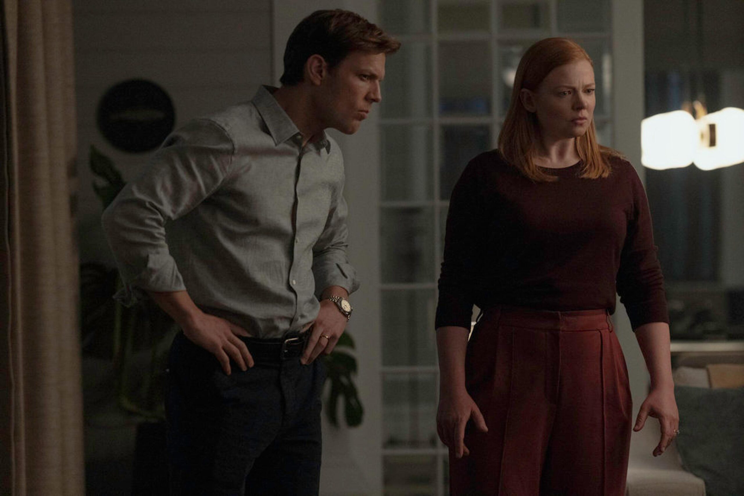 Two adults standing indoors in a dimly lit room, looking toward the right with worried expressions.