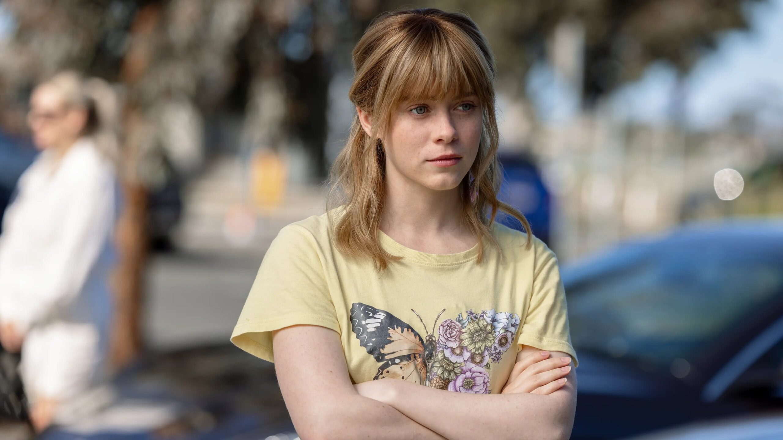 Young woman with blond hair and a yellow T-shirt stands outdoors with arms crossed, looking to the right. all her fault To jej wina