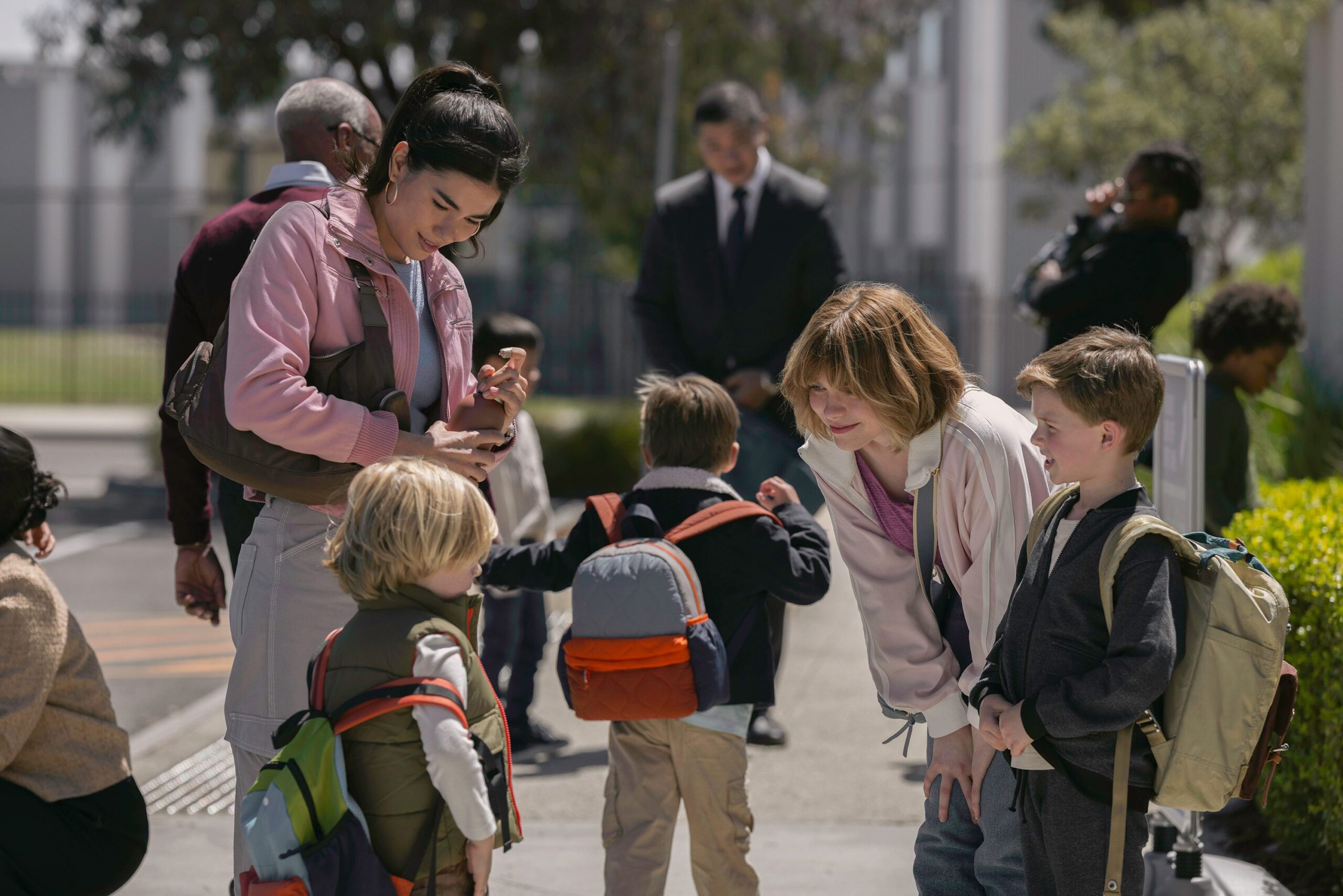 Children with backpacks gather outdoors, talking with two adults on a sunny day.