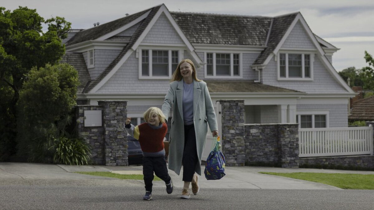 A smiling woman in a light coat walks with a young child along a suburban street in front of a large gray and white house, carrying a colorful bag. all her fault To jej wina