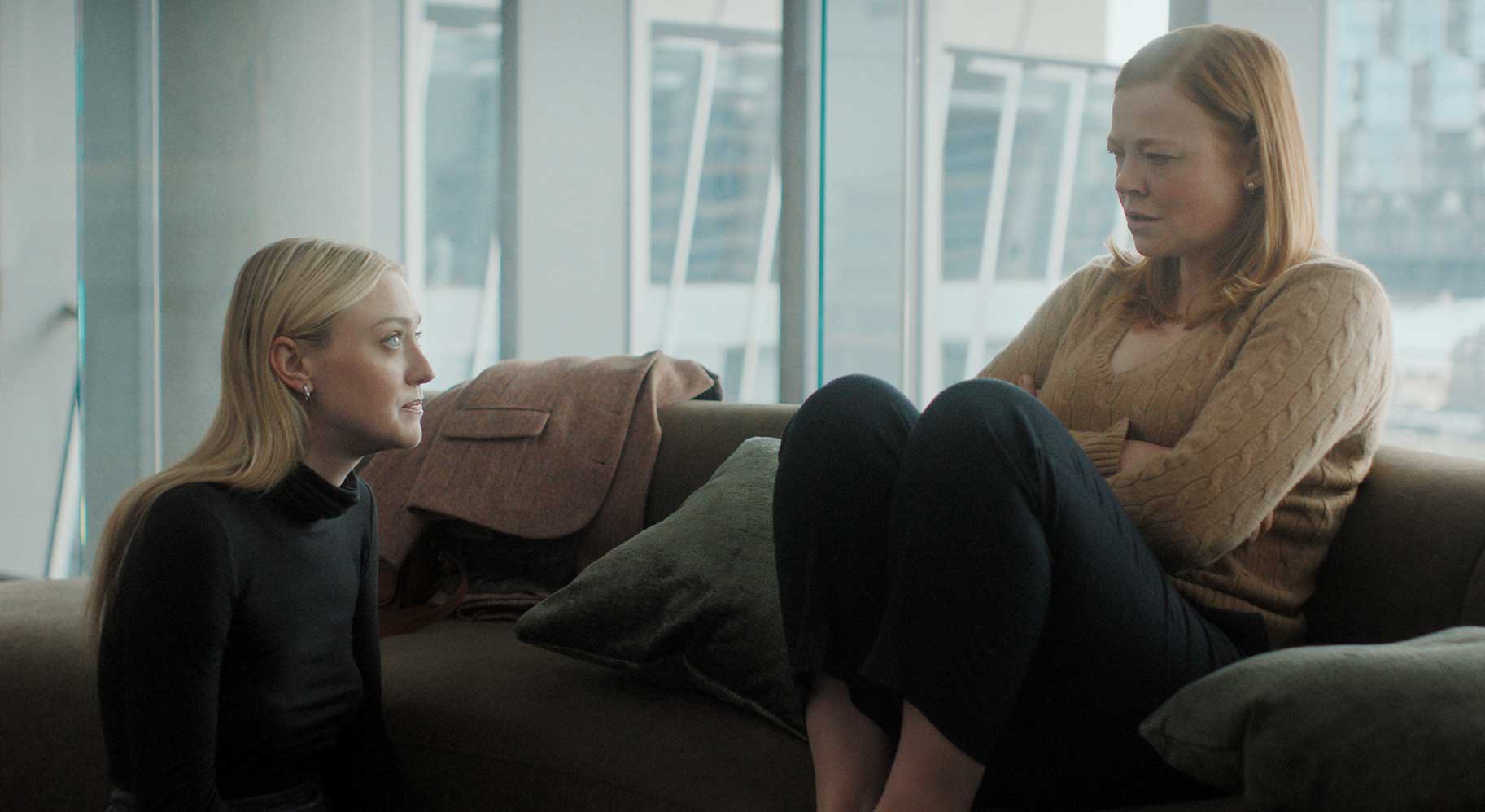 Two women sit on a couch in a modern office lounge, facing each other and talking near large windows in the background. all her fault To jej wina