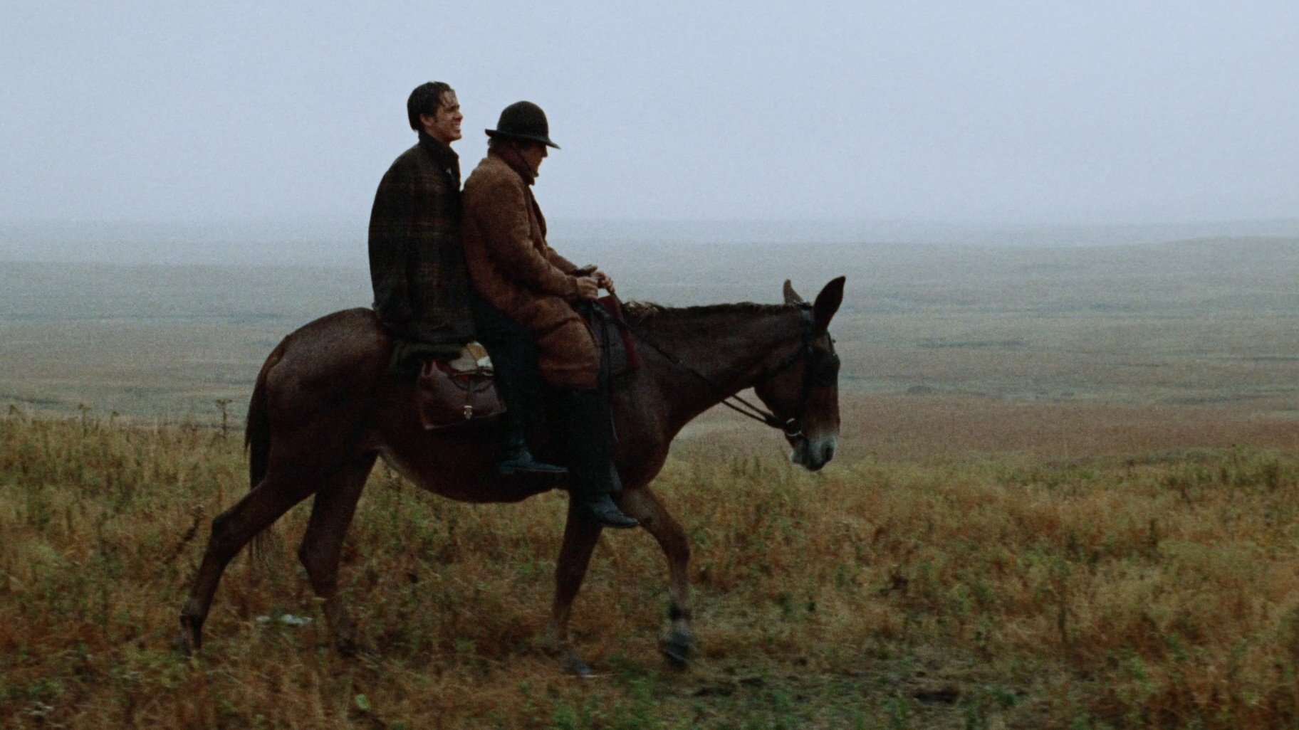 Bad Company kadr | film.org.pl Kadr z filmu Bad Company: dwóch mężczyzn jedzie na jednym koniu przez rozległy, mglisty step porośnięty suchą trawą.