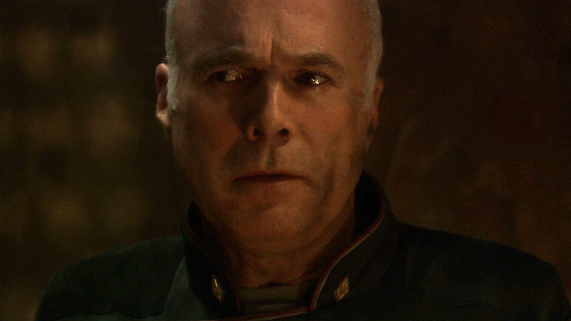 Kadr z serialu Battlestar Galactica: łysy mężczyzna w ciemnozielonym mundurze z czerwonymi lamówkami patrzy w bok z poważnym wyrazem twarzy.