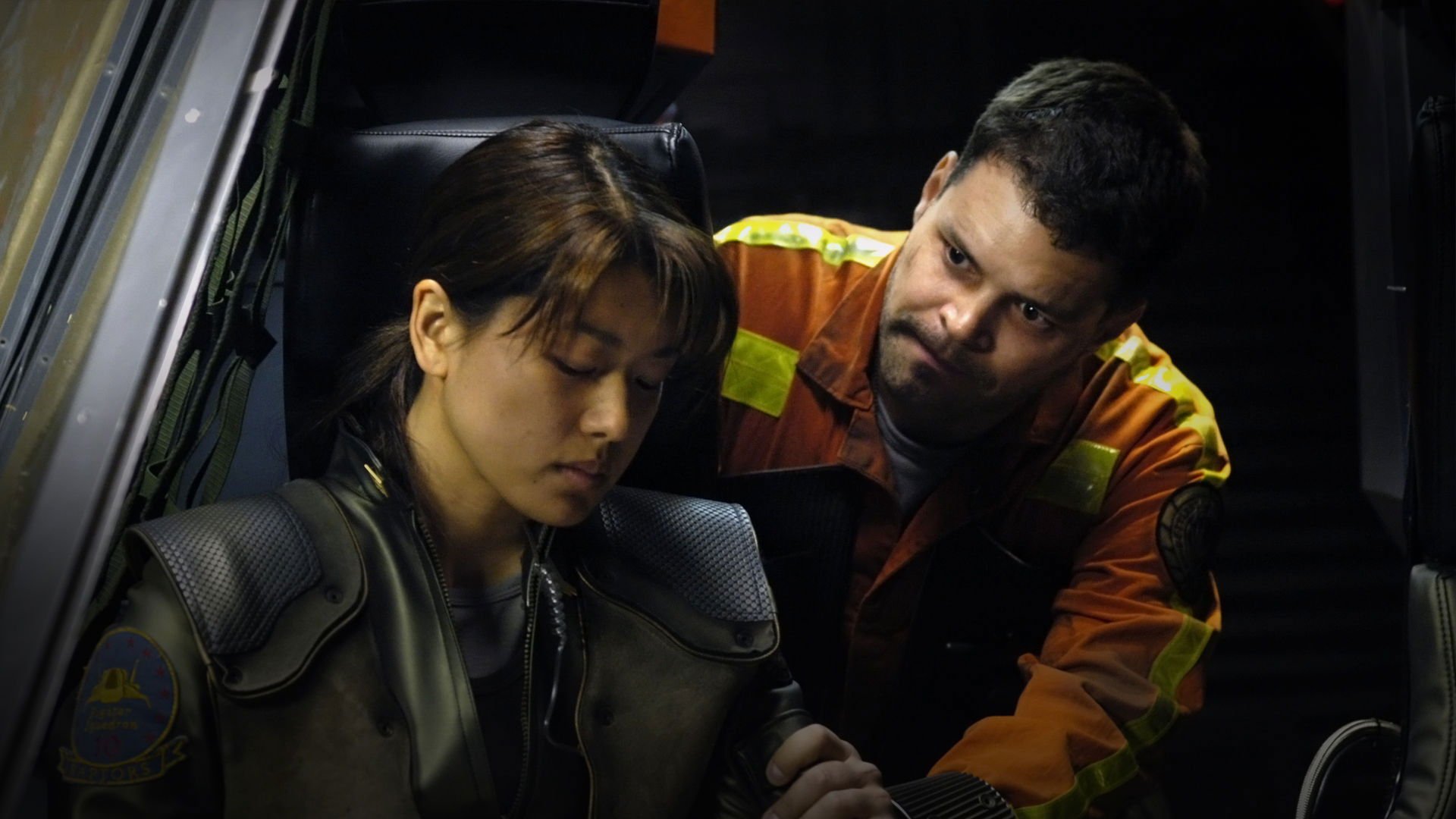 Kadr z serialu Battlestar Galactica: kobieta w ciemnej kurtce siedzi w fotelu, obok mężczyzna w pomarańczowym kombinezonie z odblaskami pochyla się nad nią