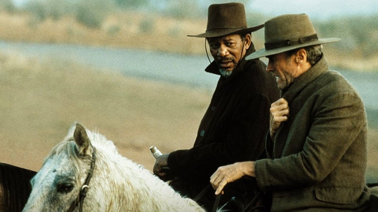 Kadr z filmu Bez przebaczenia: dwaj mężczyźni w kapeluszach jadą konno na tle suchego krajobrazu, jeden trzyma butelkę.