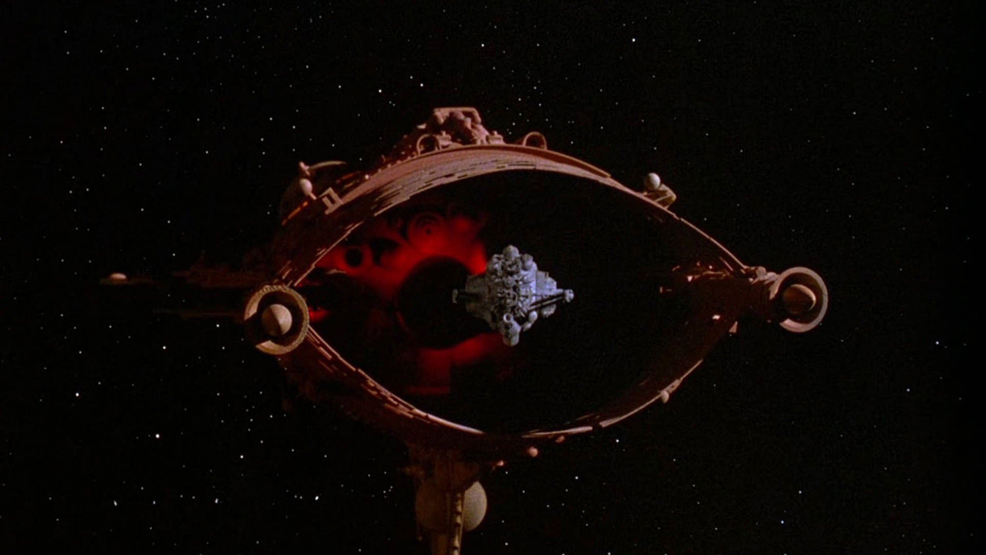 Bitwa wśród gwiazd Battle Beyond the Stars kadr | film.org.pl czerwony, owalny statek kosmiczny z obręczą i wystającymi elementami na tle gwiaździstego kosmosu, z mniejszym szarym obiektem w centrum.
