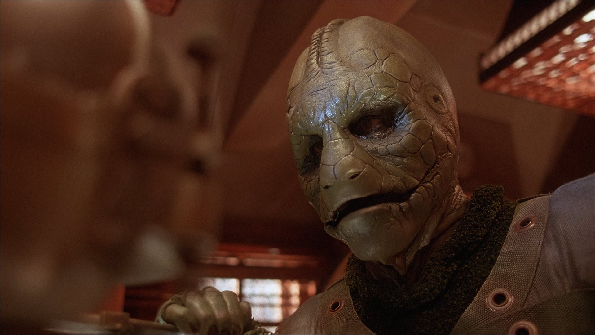 Bitwa wśród gwiazd Battle Beyond the Stars kadr | film.org.pl Kadr z filmu Bitwa wśród gwiazd: zielonoskóry humanoid z łuskowatą twarzą w ciemnym stroju, po lewej rozmyta sylwetka na pierwszym planie.