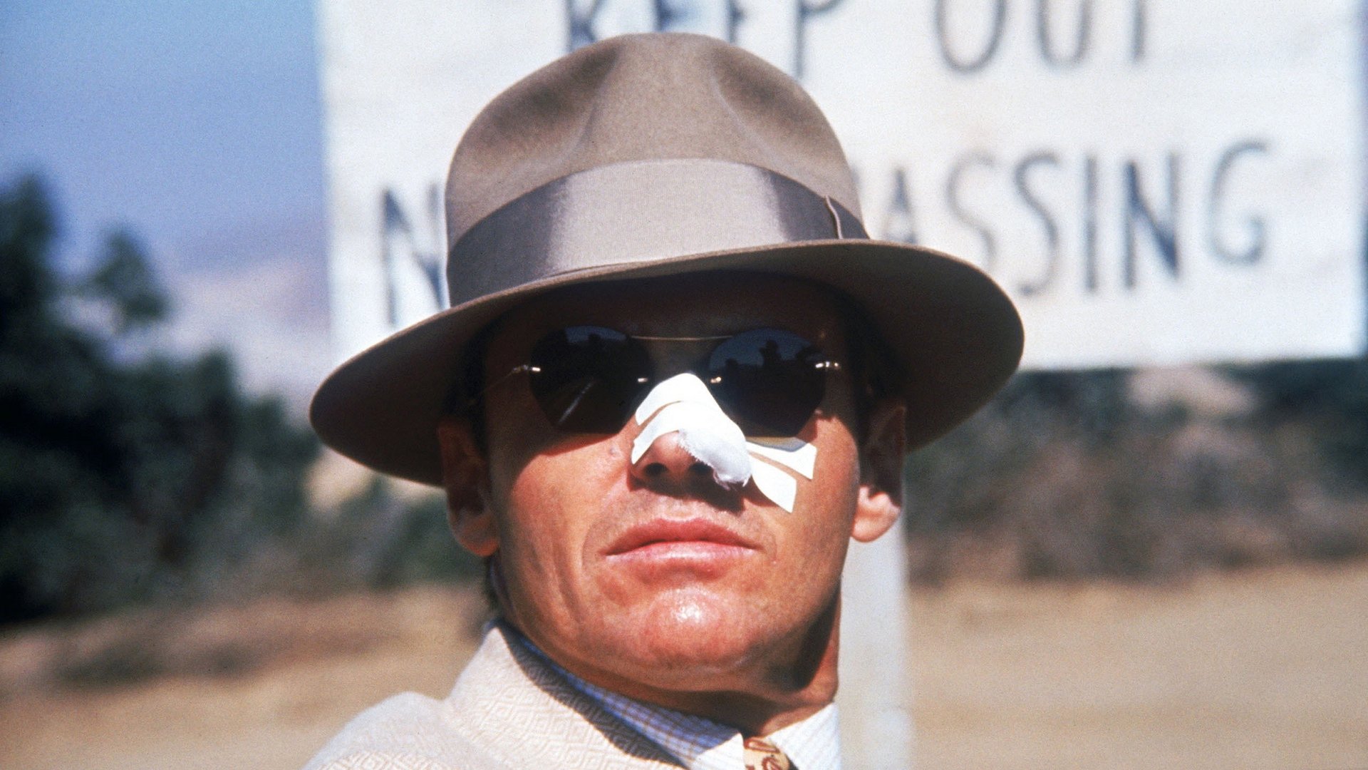 Kadr z filmu Chinatown: mężczyzna w kapeluszu, ciemnych okularach i opatrunku na nosie przy tablicy zakazu wstępu. filmy kryminalne