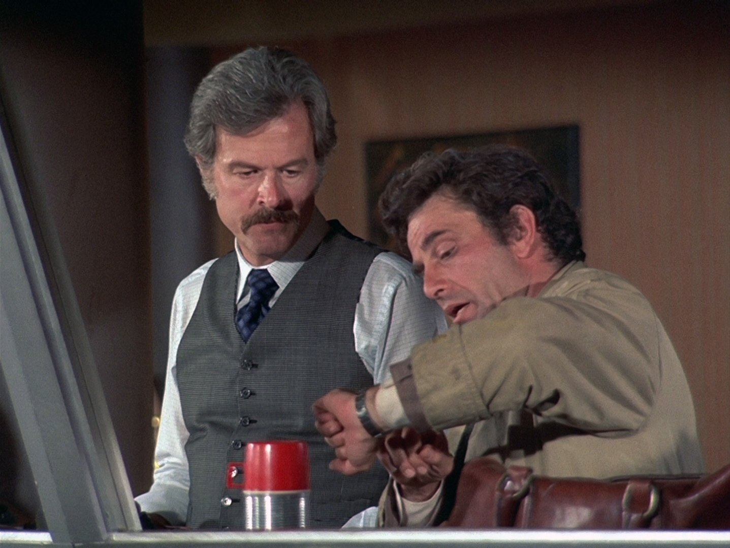 Kadr z serialu Columbo: mężczyzna w szarej kamizelce i krawacie stoi przy stole, obok drugi w beżowym płaszczu sprawdza zegarek. Sherlock Holmes