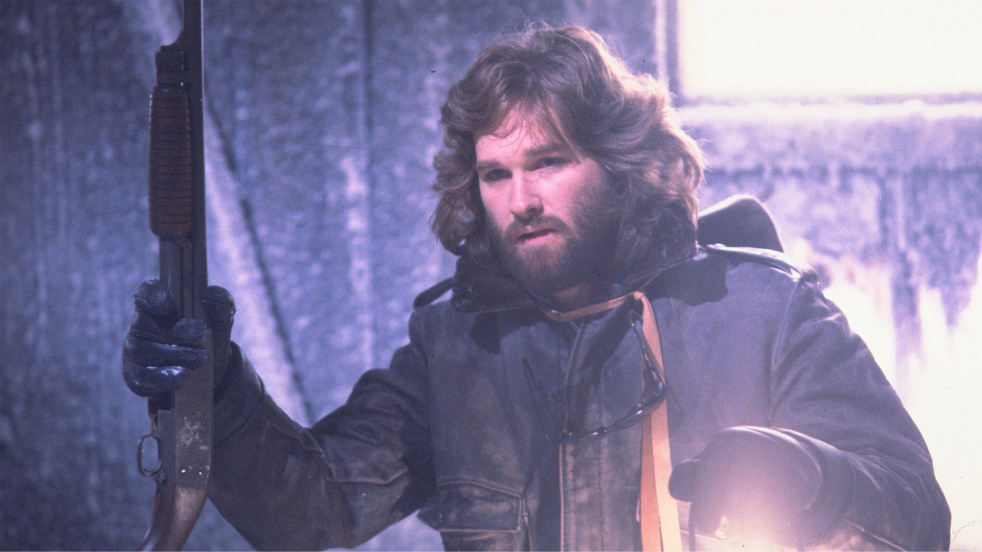 Coś The Thing kadr | film.org.pl Kadr z filmu Coś: mężczyzna z długimi włosami i brodą w skórzanej kurtce trzyma strzelbę w zimnym wnętrzu.